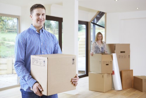A young man with short brown hair, dressed in a blue checkered shirt, is standing in a brightly lit, modern residential interior with large windows and white walls, holding a medium-sized cardboard moving box with printed labels, while smiling at the camera. In the background, a woman with long, light-colored hair, wearing a patterned blouse, is handling additional cardboard boxes of various sizes, some stacked on the floor and others being prepared for removal. The room features a wooden floor, and visible items include a roll of packing paper or bubble wrap and a few unfixed furniture pieces, indicating a house being prepared for a move in the Barnes area. The scene is illuminated by natural light coming through the glass doors, creating a clean and organized environment typical of house removals in London suburbs.
