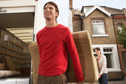 A young man with light skin, wearing a red long-sleeve shirt and brown trousers, is carrying a large beige upholstered armchair out of a white removals van parked outside a residential property in Barnes. The van door is open, revealing a glimpse of the interior with stacked cushions and packing materials. In the background, a woman with dark hair, dressed in a white top with grey stripes, is standing near the house's brick exterior, which features a bay window and a white front door. The scene takes place on a cloudy day on a quiet suburban street with similar brick houses. The young man appears to be smiling slightly as he relocates the furniture, and the environment suggests an ongoing house removal or relocation arranged by Barnes Man with Van, part of professional house removals services serving the Barnes and surrounding areas.