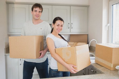 A young man and woman, both with light skin and casual clothing, standing inside a modern kitchen with light grey cabinets and a white tiled backsplash, are holding cardboard boxes used for moving house. The woman, smiling, is positioned in the foreground, holding a medium-sized box with both hands, while the man, standing slightly behind her, is carrying a larger box. To the right, on the kitchen countertop, there is a stack of unopened boxes, and a white sink is visible beneath a large window that allows natural light to illuminate the scene. The background shows a neutral-toned interior typical of a residential property, suggesting a home environment in a suburban area such as Barnes or nearby in southwest London. The scene captures the process of packing or unpacking during a house removal, with the visible items made of cardboard, and the setting indicative of professional house removals services provided by Barnes Man with Van.